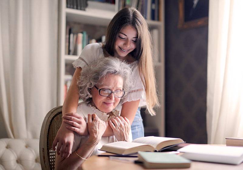 A young girl hugging an elderly woman while she reads a book at a table