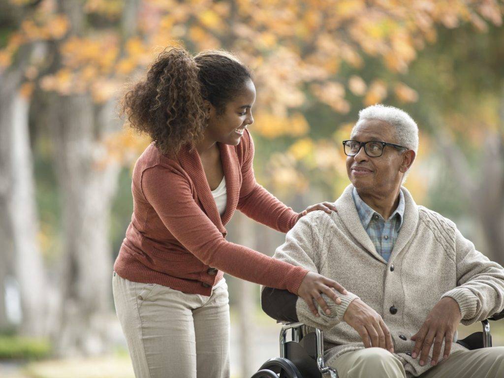 An elderly man sits in a wheelchair while a young woman pushes him around outside, both smiling