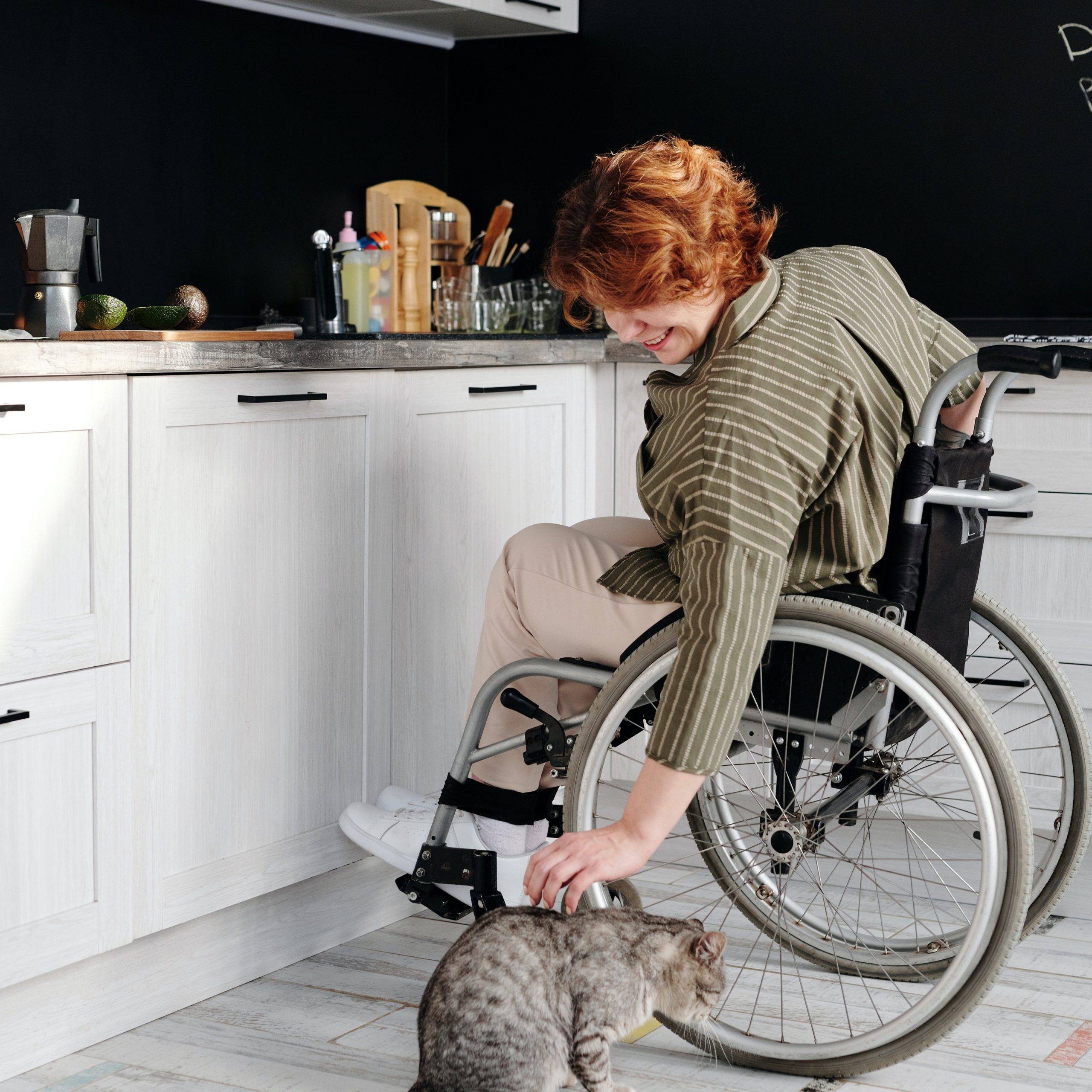 Woman in wheelchair in her kitchen, petting a cat on the floor