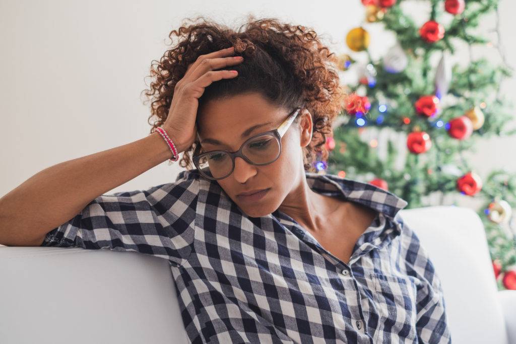 woman sits with head in hands on couch, christmas tree in background