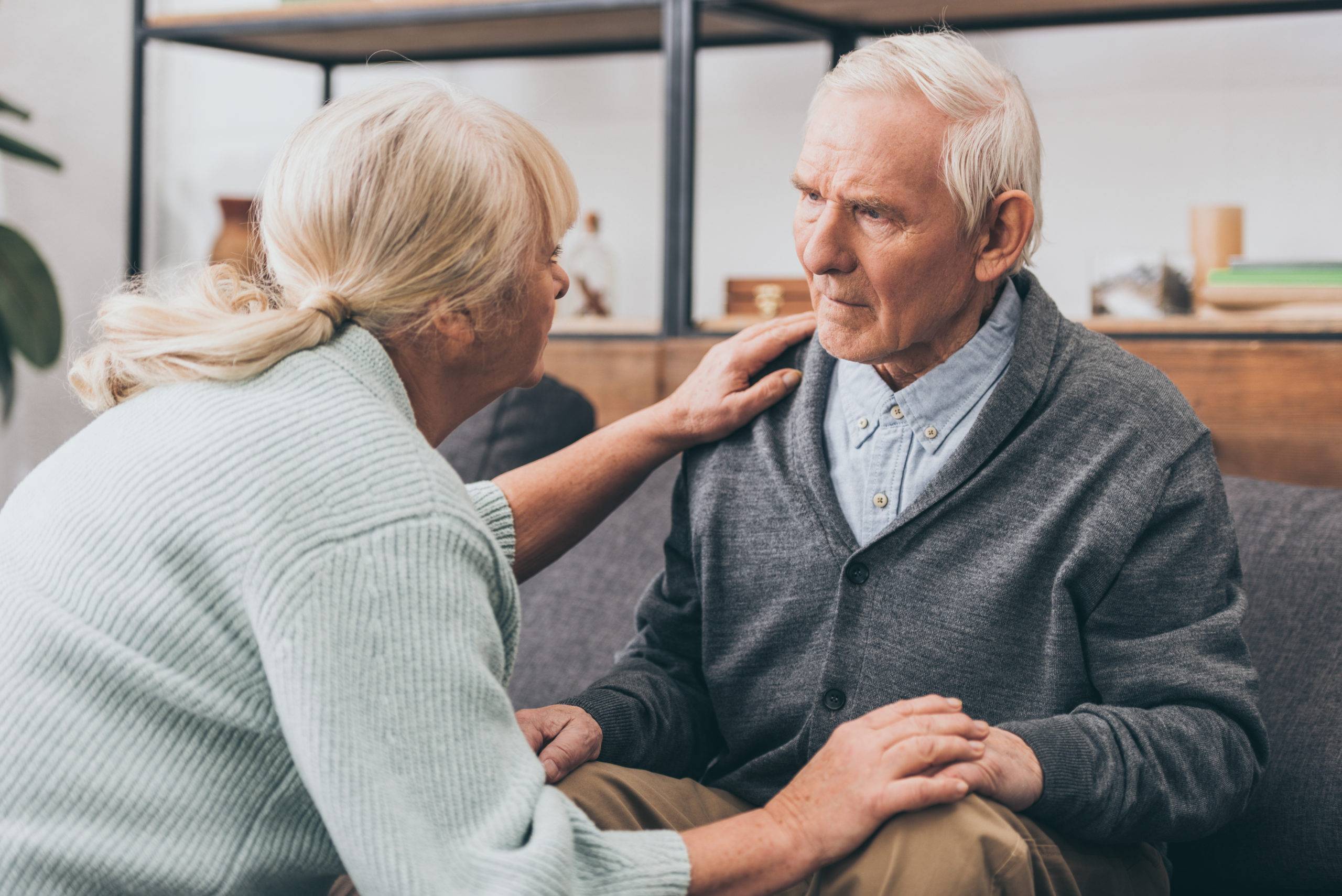 Elderly man on couch with concerned look, elderly woman holding his hand and shoulder