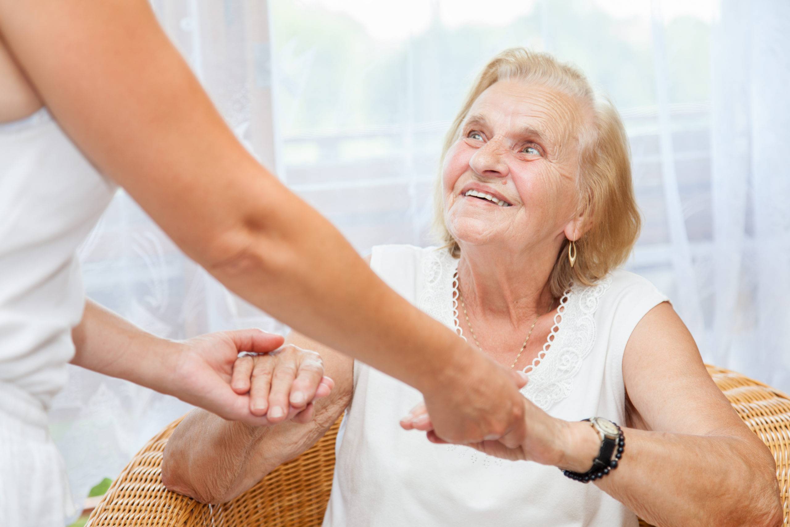 elderly woman seated in chair, receiving support to stand from someone