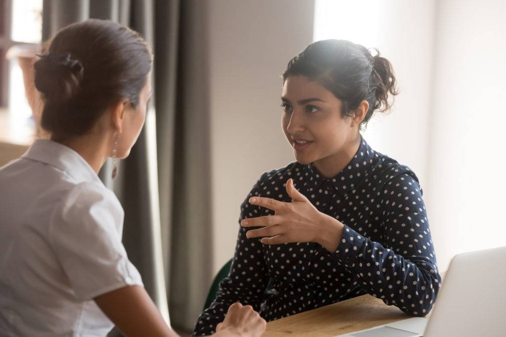 Two adult woman sitting at desk, having serious conversation