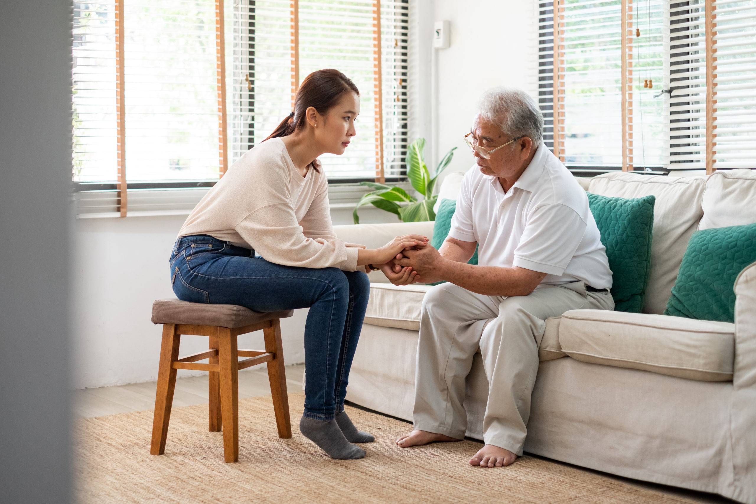 Elderly man sits on couch, adult daughter sits on stool and holds hands