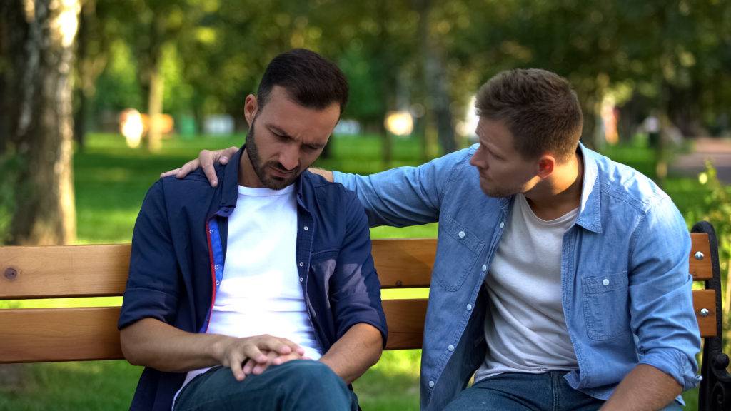 Young male sits on bench outside, friend sits next to and consoles him