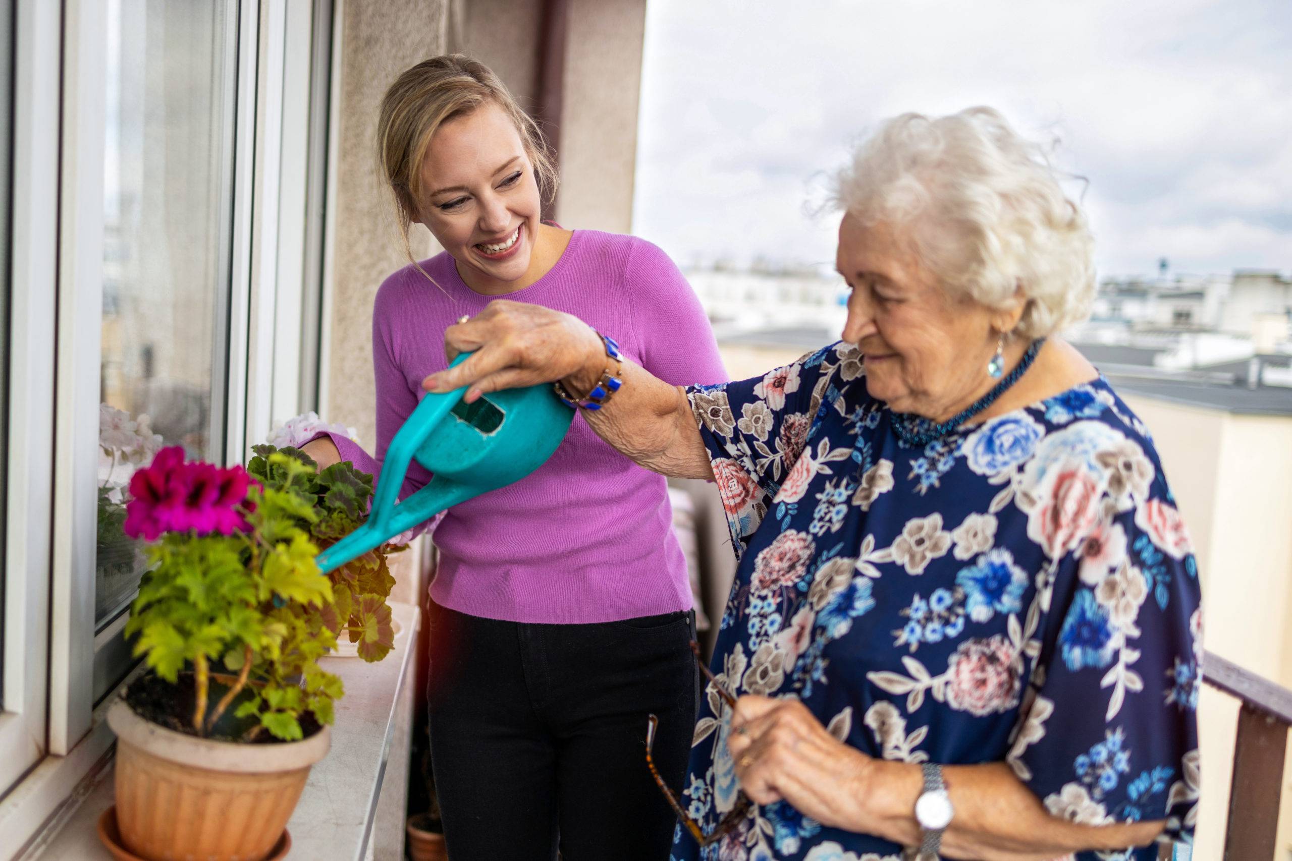 Elderly woman waters plants on balcony, young woman smiling next to her