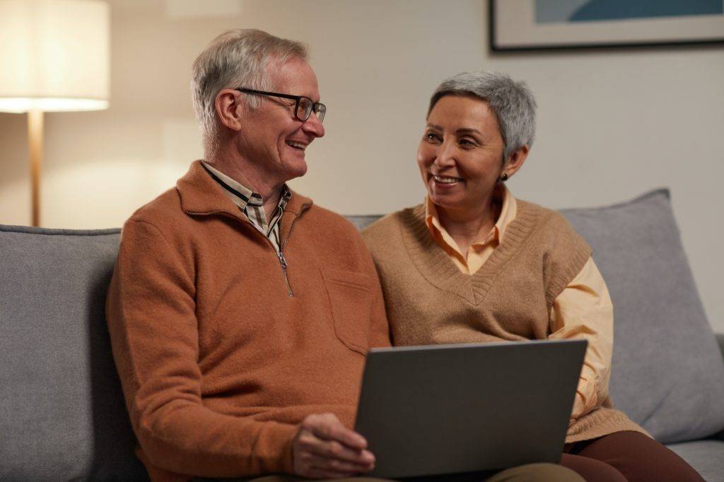 Elderly couple sits on couch using laptop and smiling