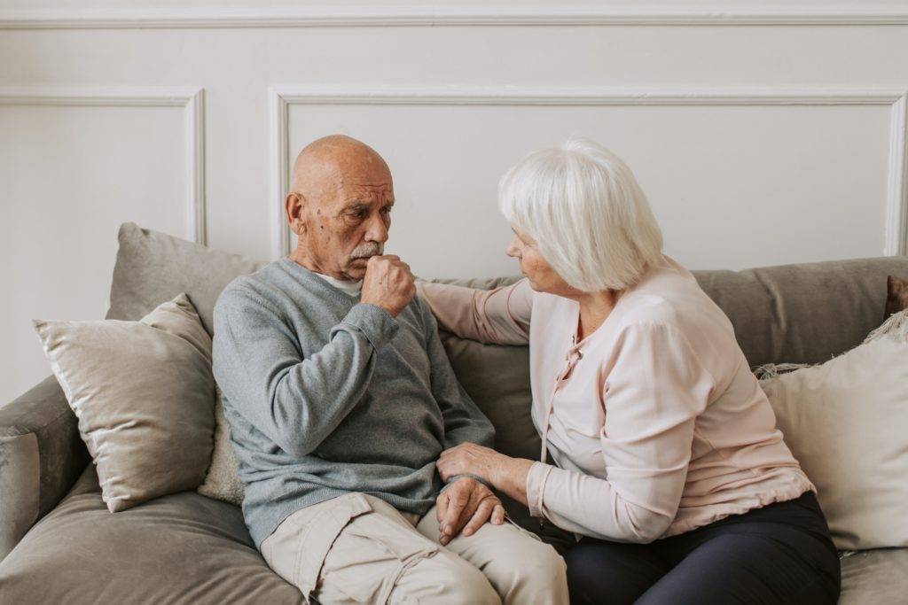 Elderly man sits on couch with elderly woman, distressed
