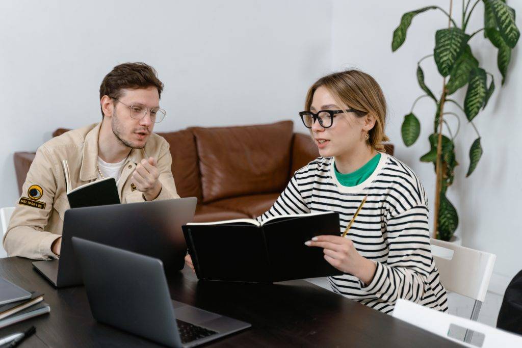 Mlilenial woman and man seated at table using laptops and journals