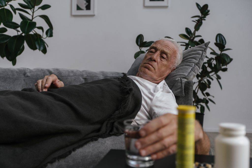 Elderly man laying on couch with blanket, holding glass of water