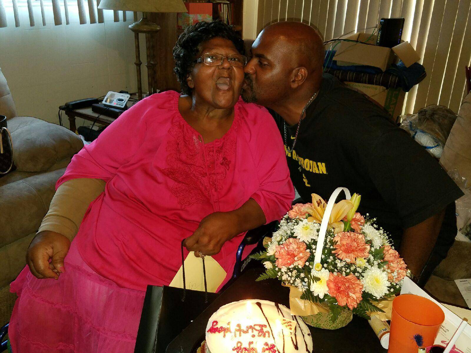 Adult man kissing elderly mother on cheek, seated in front of birthday cake
