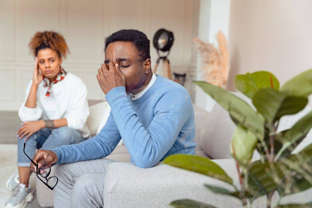 Adult man sitting on couch, holding nose in distress. Adult woman sits in background