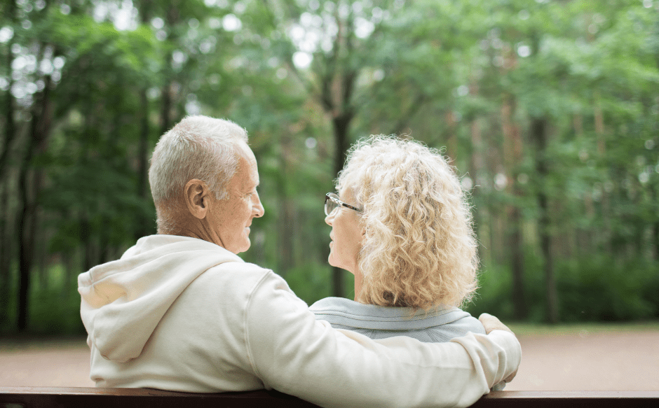 image shows two adults sitting on a park bench gazing at each other. the image is shot from behind them