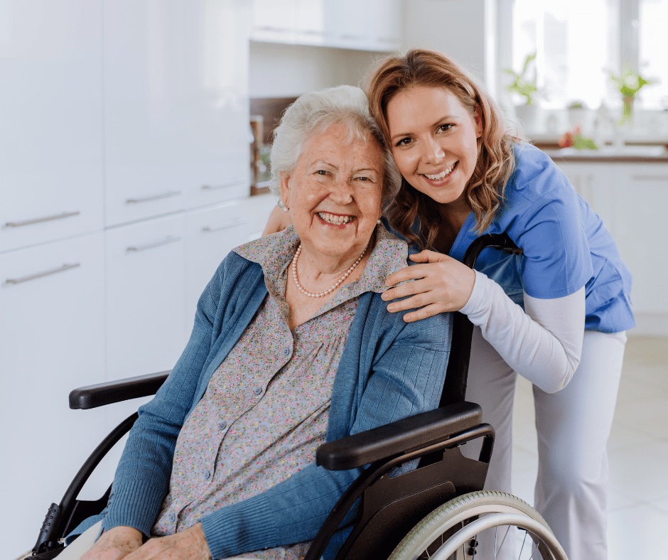 Image shows an elderly woman in a wheel chair being hugged by a younger woman in scrubs, both are smiling at the camera