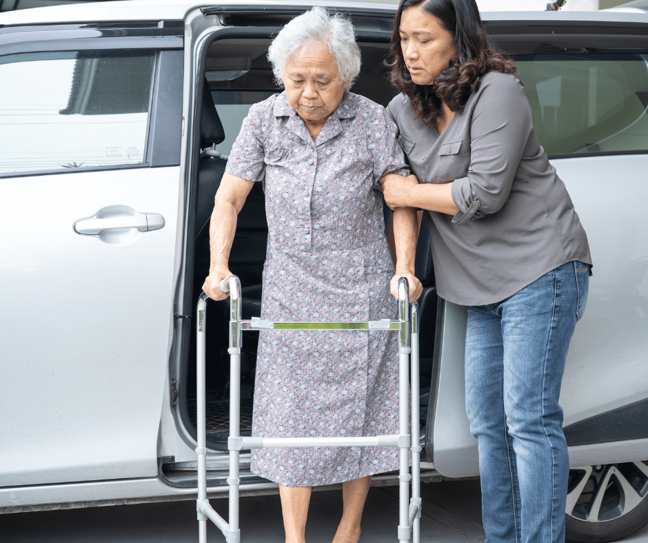 a younger woman helps an older woman with a walker out of a van.