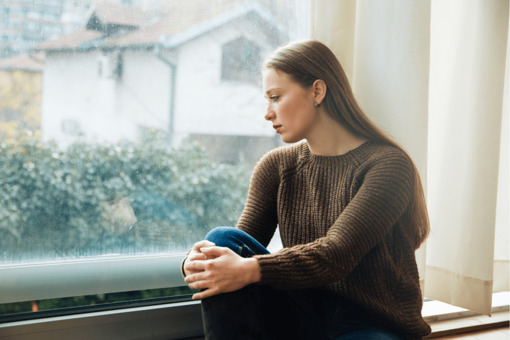 A young woman sitting near a window looking outside, with a expression of guilt